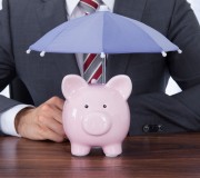 Businessman Sheltering Piggybank With Umbrella At Desk